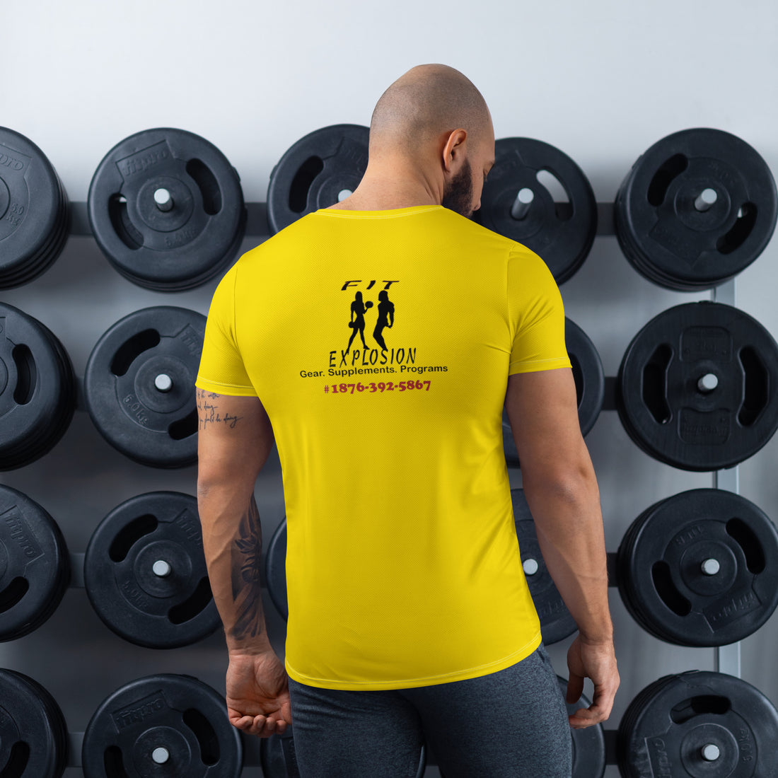 A male fitness professional wearing a custom yellow branded t-shirt featuring the 'Fit Explosion' logo, phone number, and services including gear and supplements, standing in front of a gym weight rack.