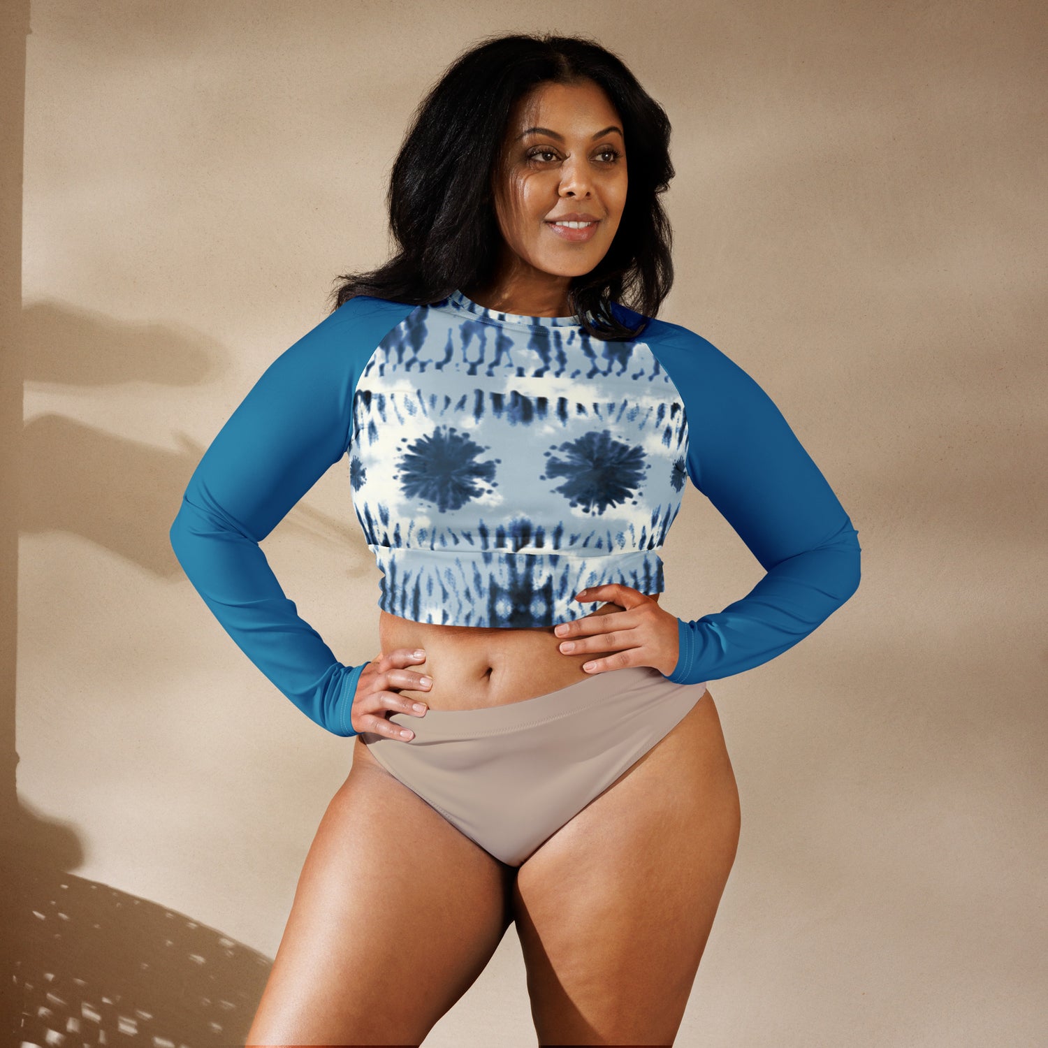 Image all-over-print-recycled-long-sleeve-crop-top-white-front-6a11f00543687.jpg: A smiling woman wearing IslandWear's high-performance blue tie-dye long sleeve crop top rash guard with solid blue raglan sleeves against a neutral sandy backdrop.