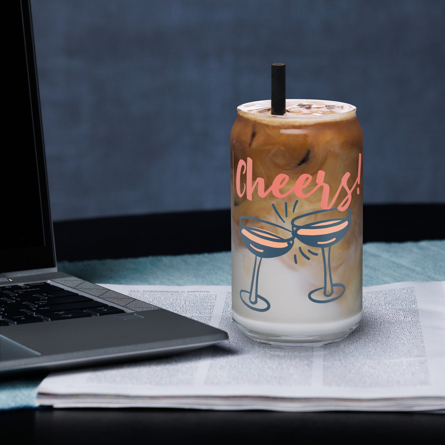 Glass tumbler with 'Cheers!' text and drink design next to a laptop on a desk.