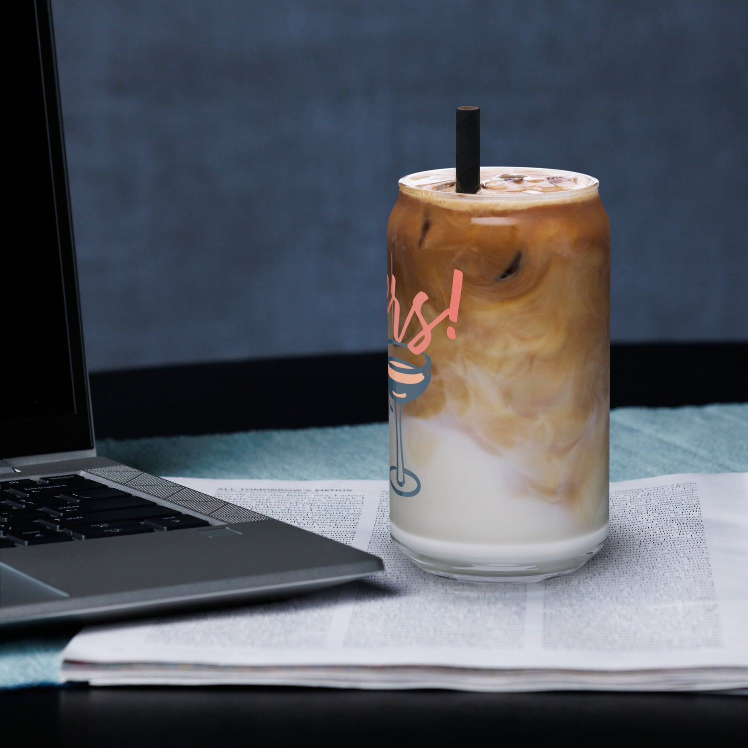 Iced coffee in a glass with a straw next to a laptop on a desk.