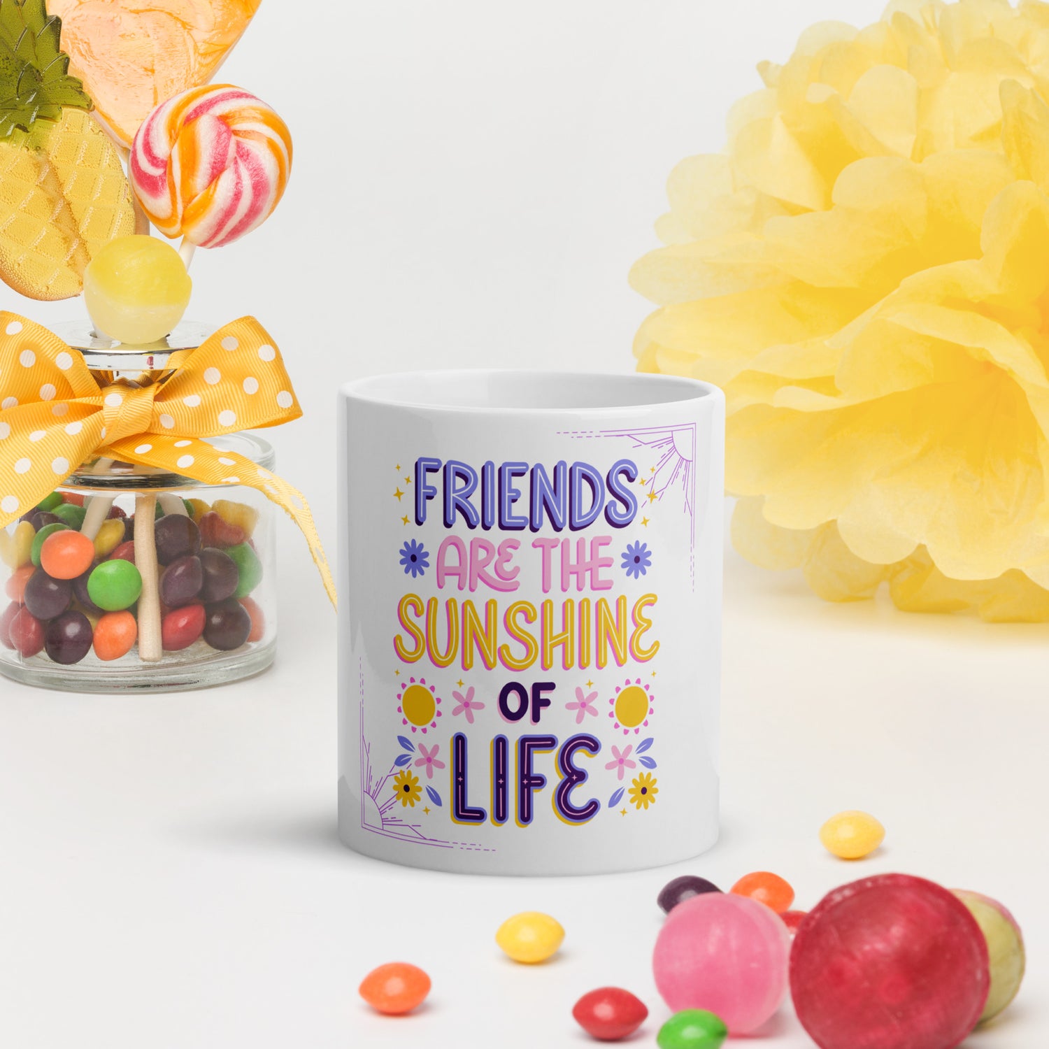 White ceramic coffee mug with "Friends are the Sunshine of Life" quote, surrounded by colorful candy and a yellow bow.